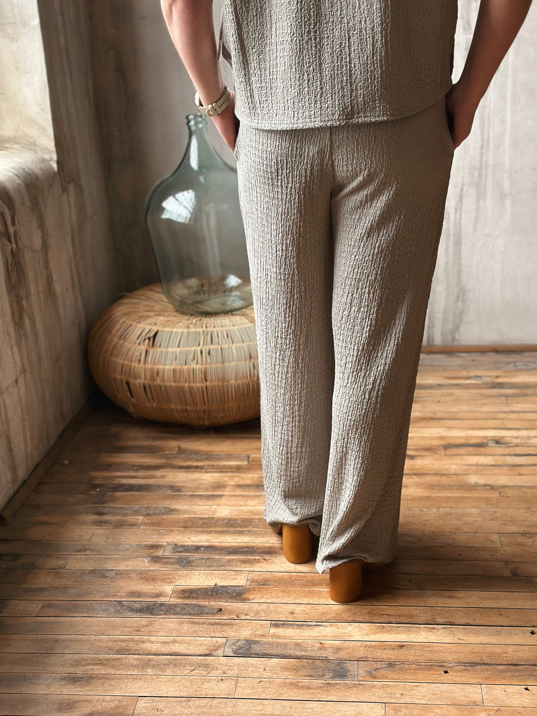 A person wearing a sage textured outfit standing on a wooden floor with a large glass vase in the background.