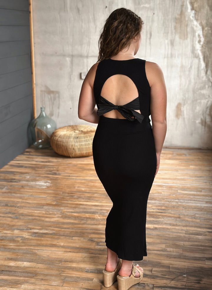 Woman wearing a black dress with an open back and a bow in a room with wooden floor and textured wall.