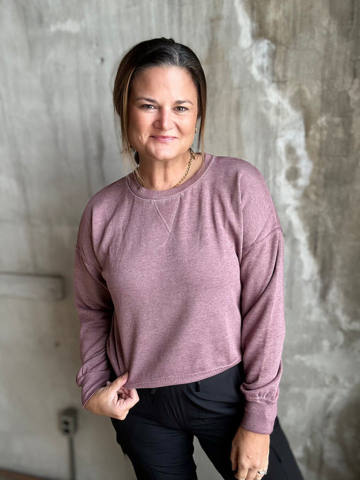 A woman wearing a dusty rose long-sleeve crewneck sweatshirt and black pants, standing against a textured concrete wall.