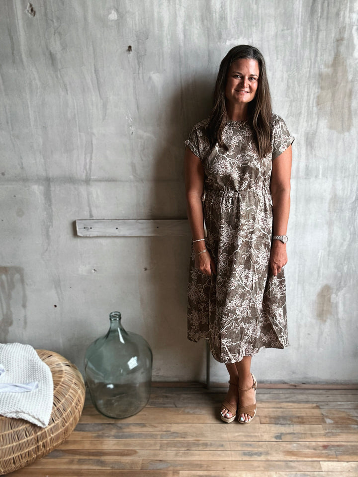 Woman in a floral dress standing against a textured wall with a large glass container and woven basket on the floor.