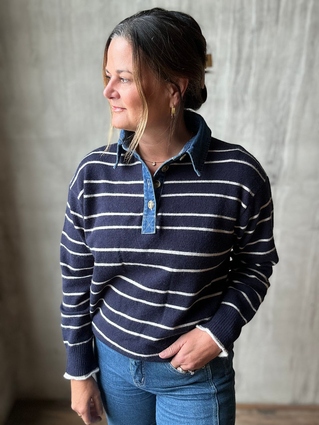 A woman wearing a navy blue and white striped knit polo sweater with a light blue denim collar and button placket, paired with blue jeans, standing against a textured gray wall.