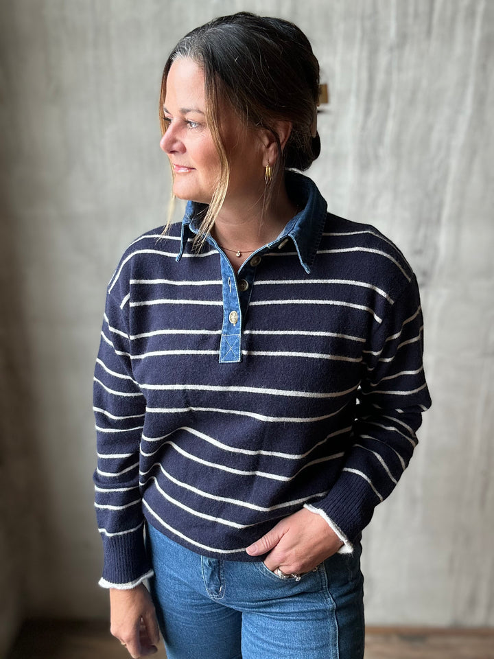 A woman wearing a navy blue and white striped knit polo sweater with a light blue denim collar and button placket, paired with blue jeans, standing against a textured gray wall.