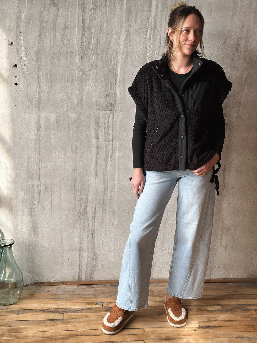 Woman wearing a black quilted jacket, light blue jeans, and brown shoes against a gray wall.