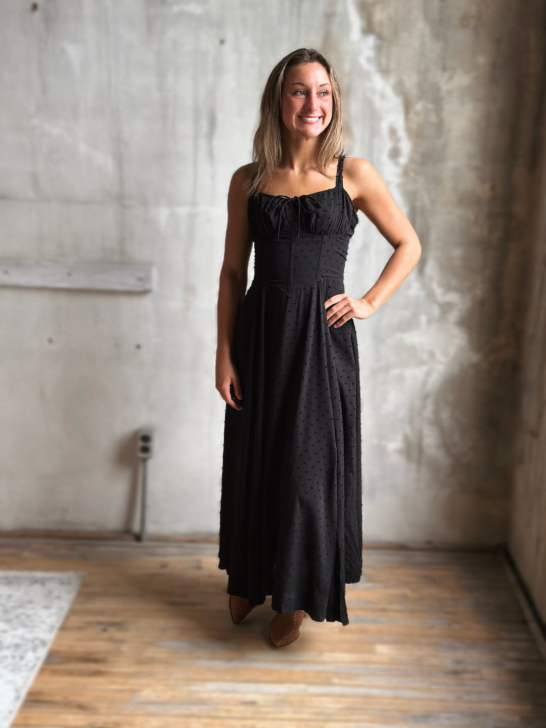 Woman wearing a black dress standing in a room with concrete walls and wooden floor.