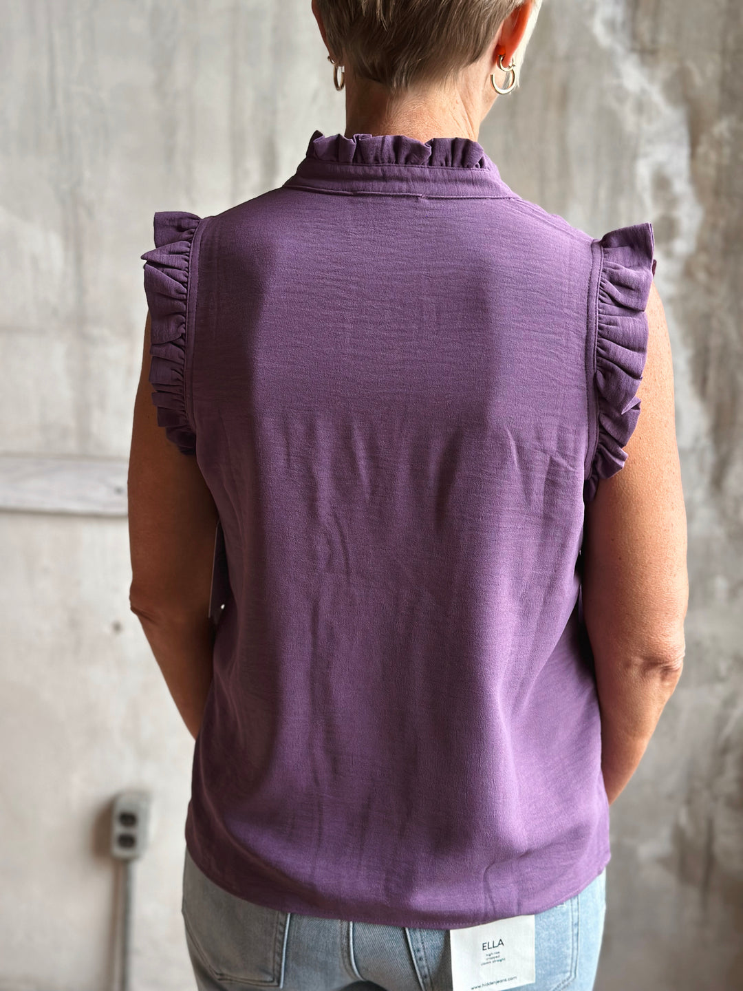 Person wearing a purple sleeveless top with ruffled shoulders against a gray wall.