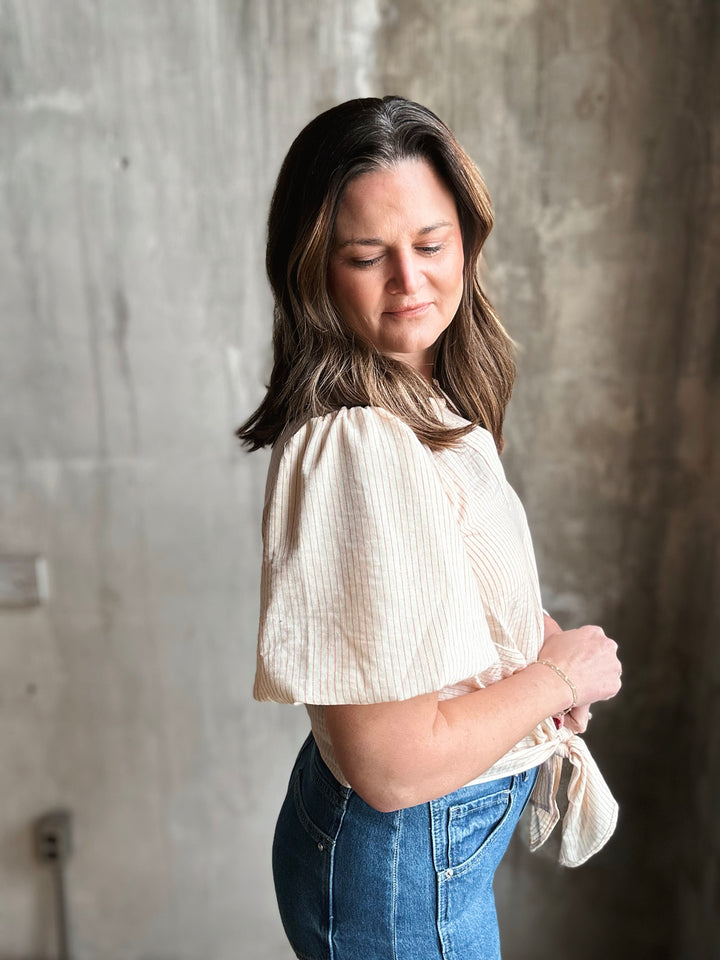 Woman wearing a beige blouse and blue jeans against a concrete wall.