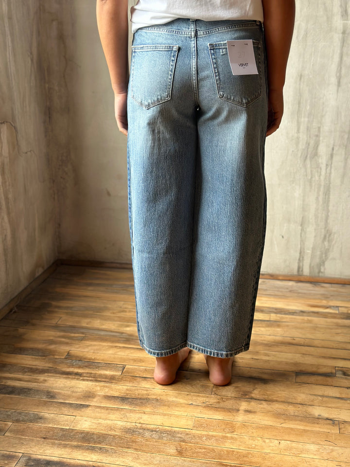 A person shown from the back wearing light-wash wide-leg denim jeans, standing barefoot on a wooden floor against a textured concrete wall.