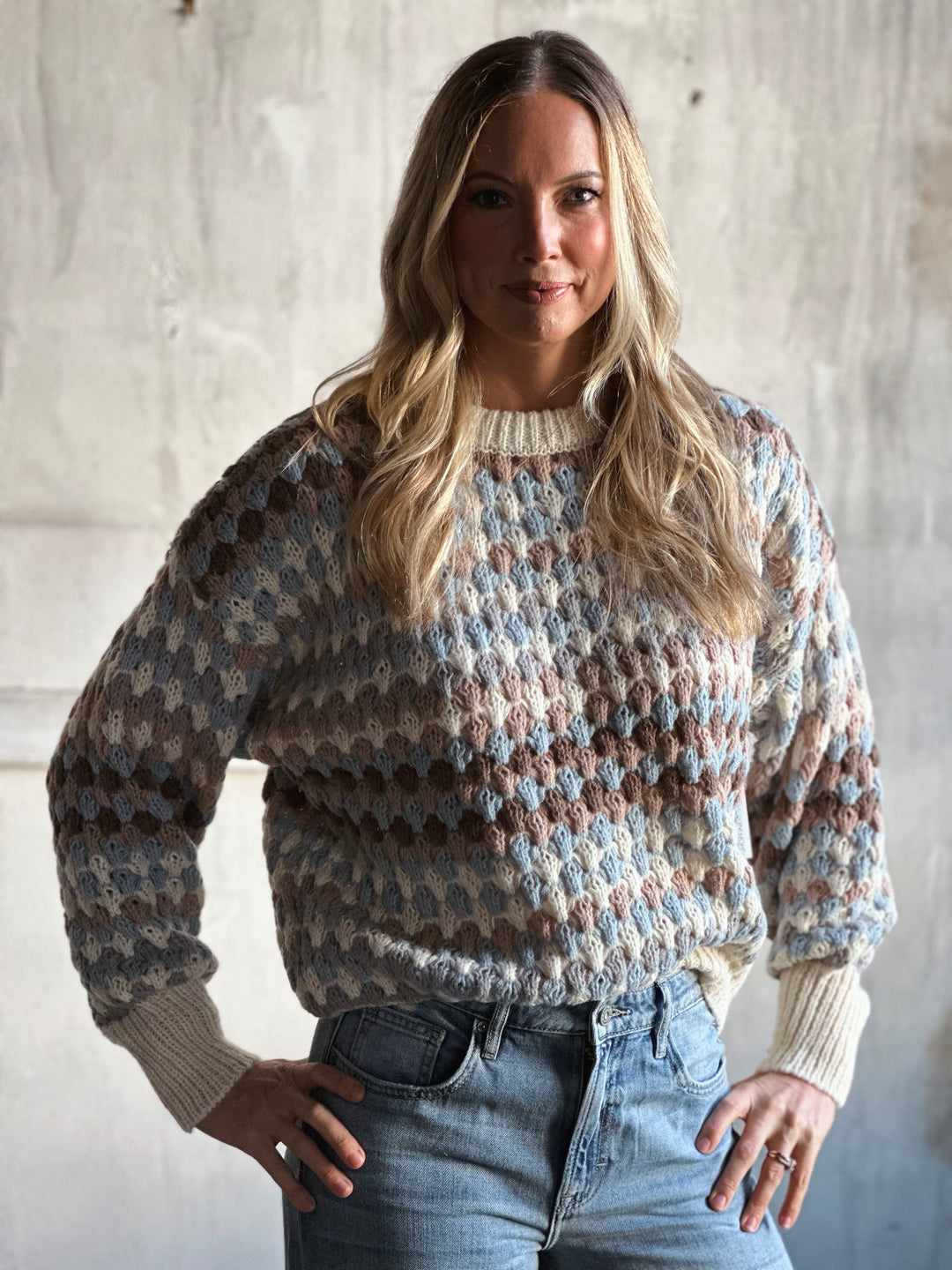 Woman wearing a patterned sweater and jeans against a concrete wall.