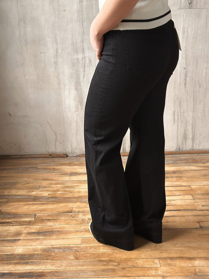 Person wearing black wide-leg jeans standing on a wooden floor with a concrete wall background