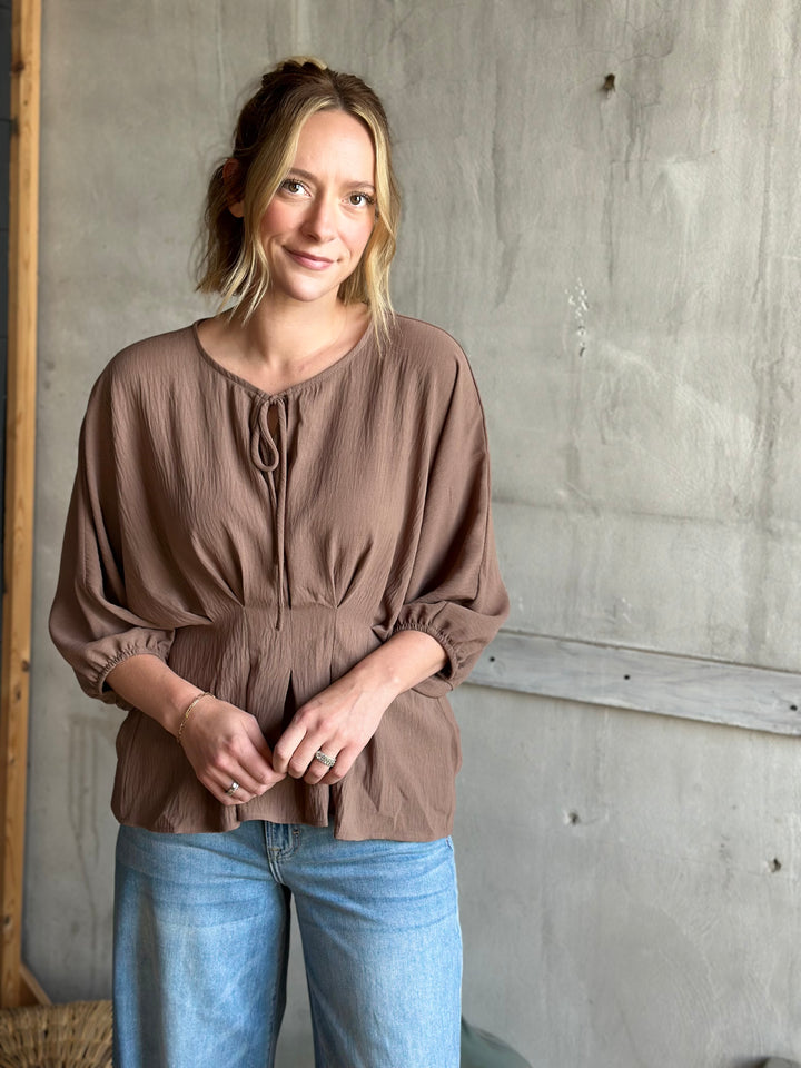 Woman wearing a brown blouse and blue jeans standing against a concrete wall.