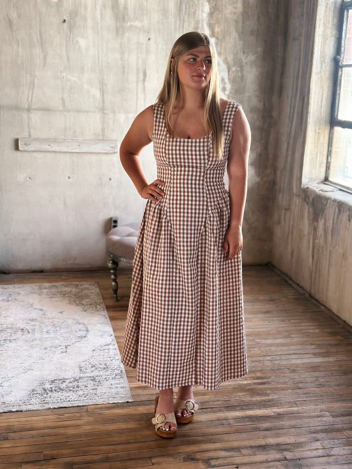 Woman wearing a brown and white plaid dress standing in a room with wooden floors and concrete walls.