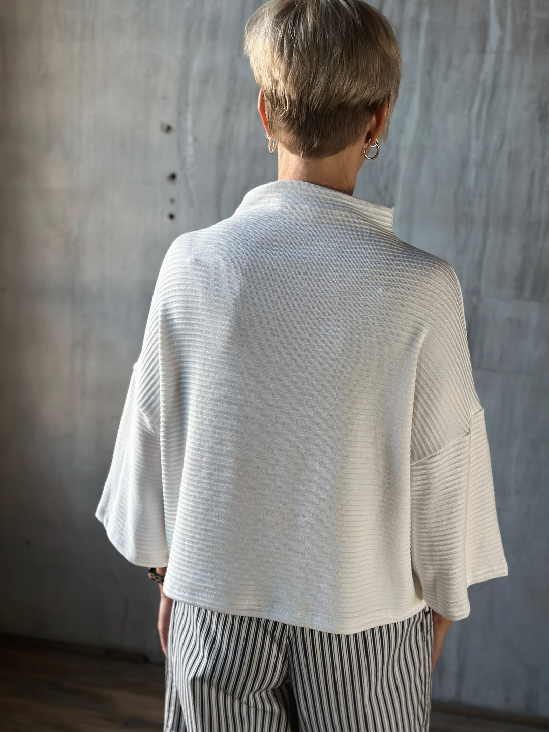 Person wearing a light-colored cardigan and striped pants against a textured wall.