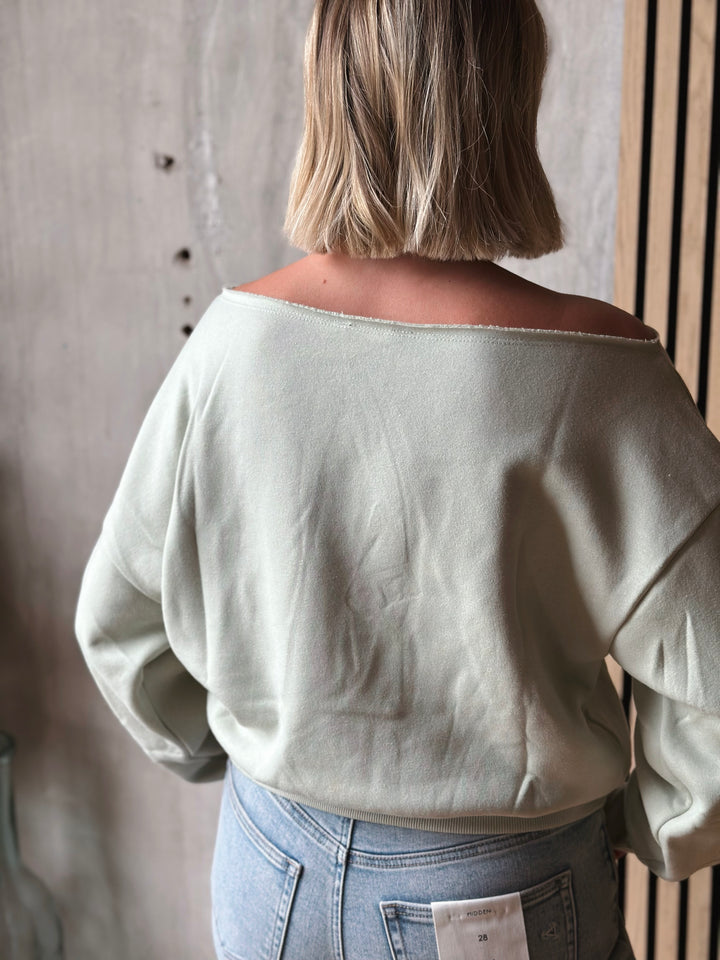 Person wearing a light green off-shoulder top and blue jeans against a neutral background
