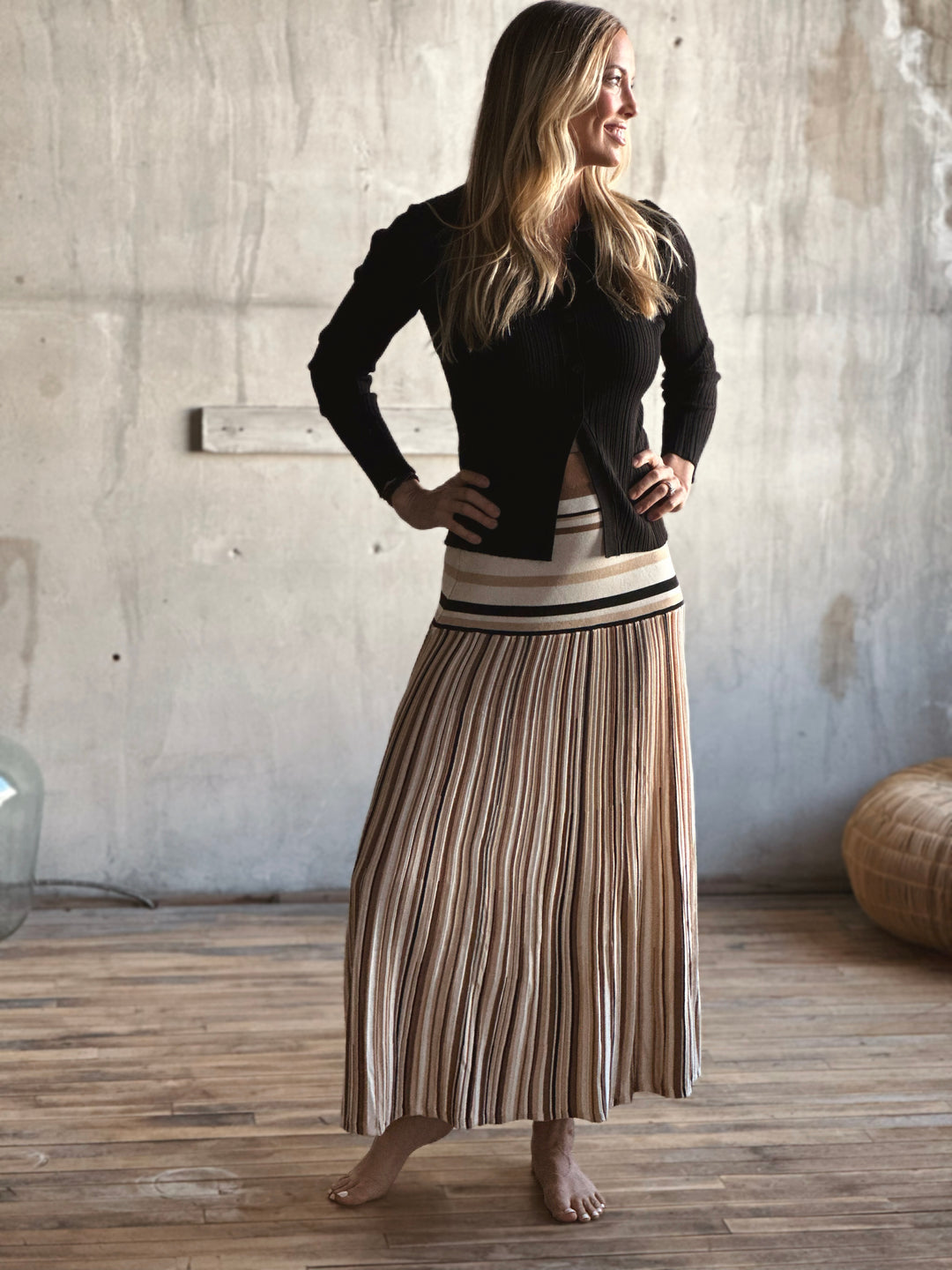 Woman wearing a black cardigan and striped skirt standing in a room with wooden floor and concrete wall.