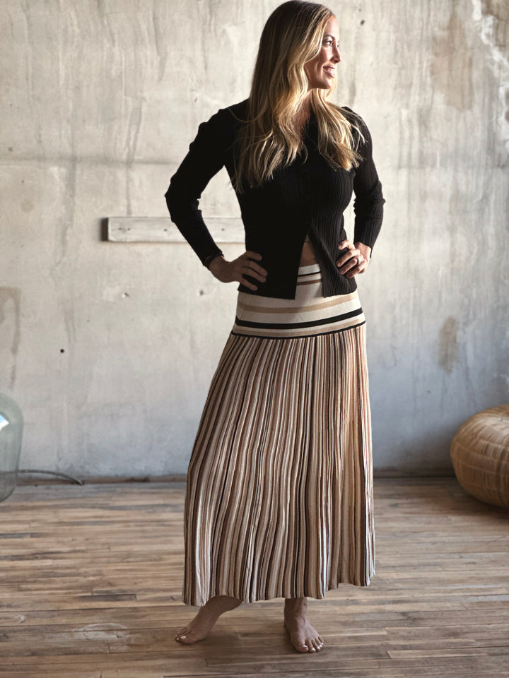 Woman wearing a black cardigan and striped skirt standing in a room with wooden floor and concrete wall.