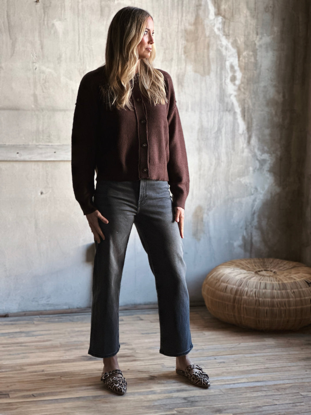 Woman wearing a brown cardigan and gray pants standing against a textured wall.