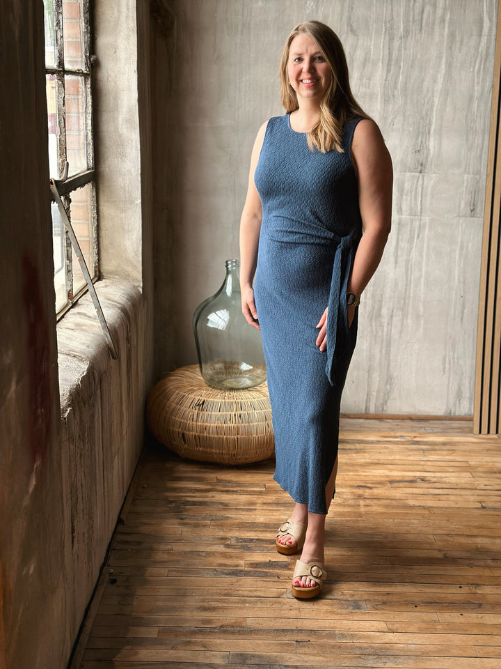 Woman in a blue dress standing in a room with wooden floor and textured walls.