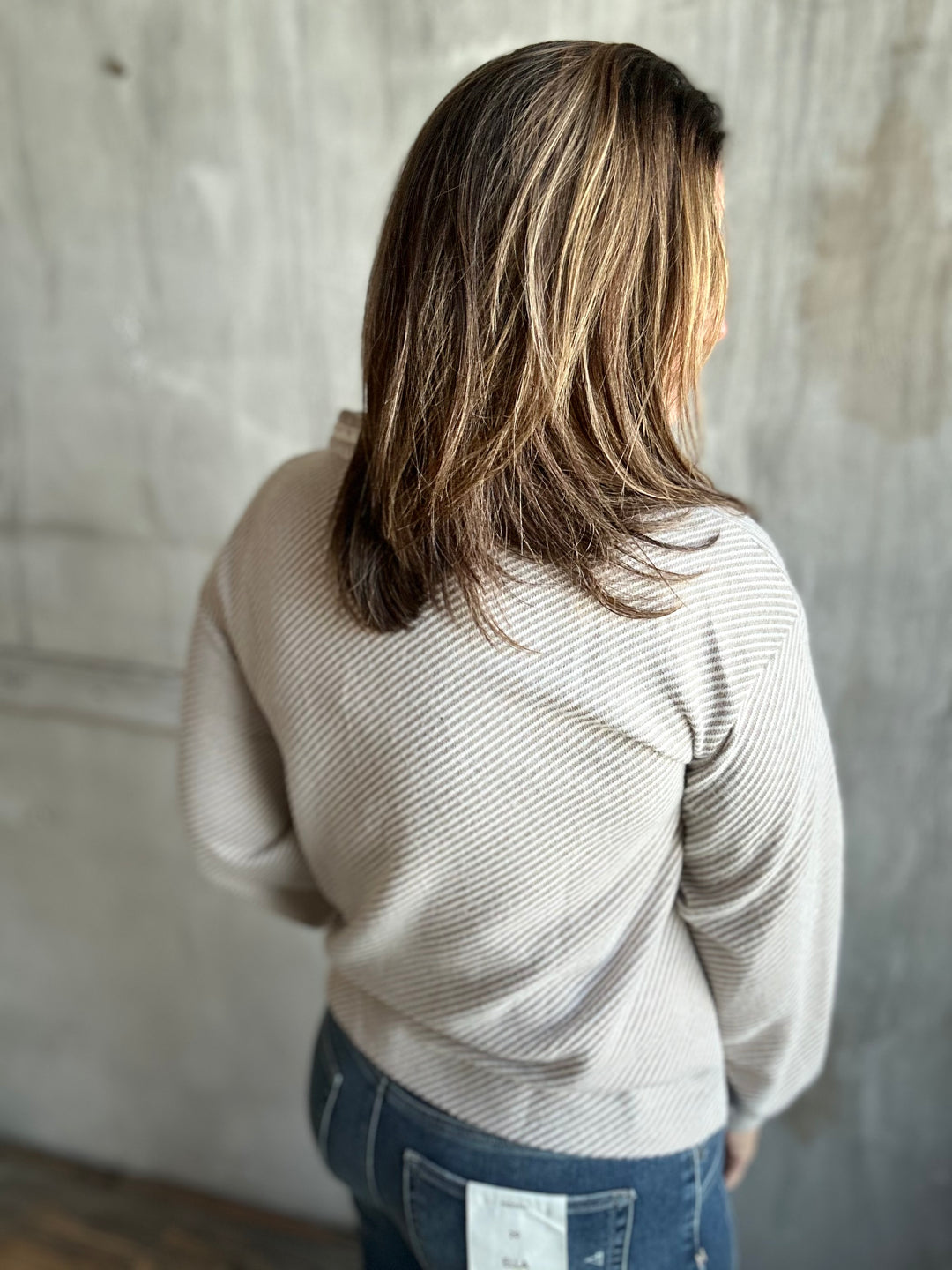 A woman viewed from behind wearing a beige ribbed knit sweater and blue jeans, standing against a textured concrete wall.