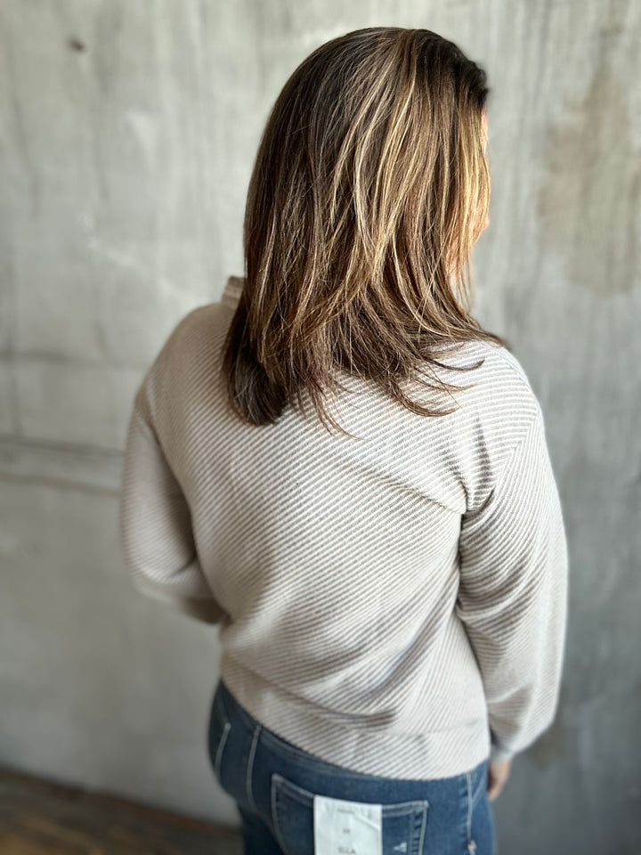 A woman viewed from behind wearing a beige ribbed knit sweater and blue jeans, standing against a textured concrete wall.