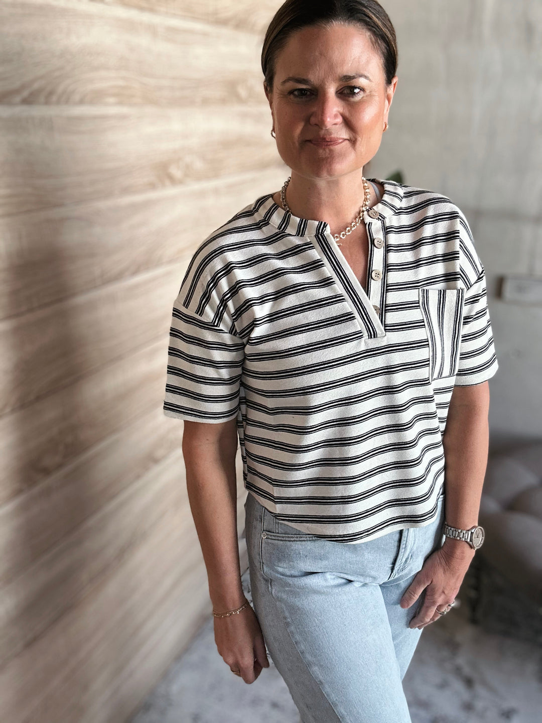 A woman wearing a short sleeved black and white striped v-neck shirt with buttons. With a pocket on the left side of the shirt. 