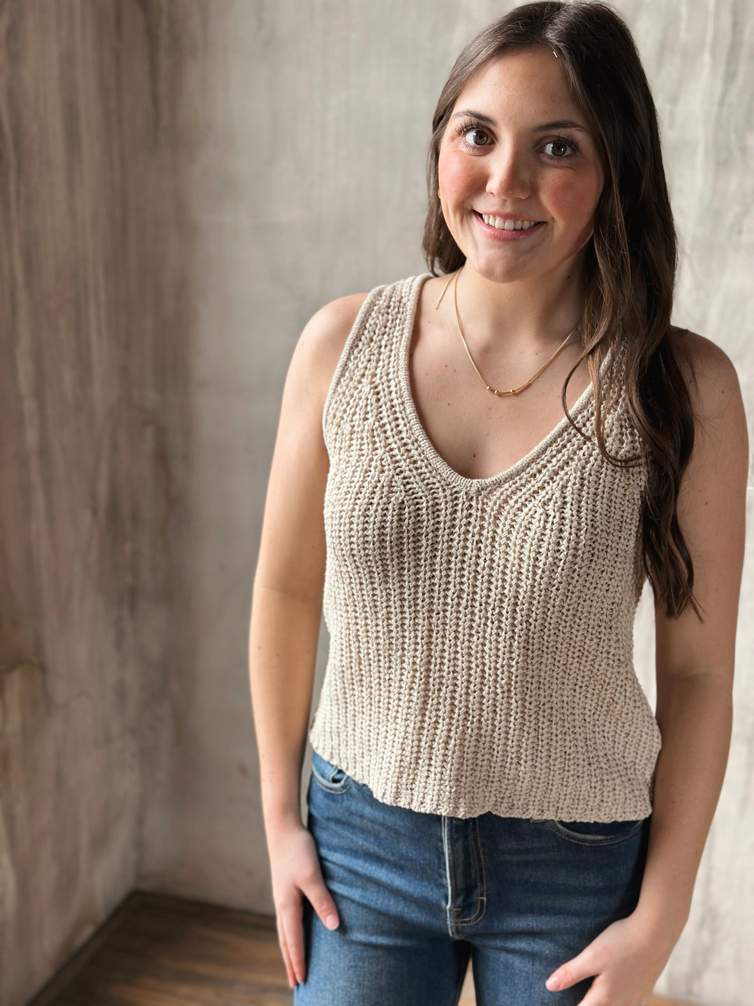 Woman wearing a beige knitted tank top and blue jeans against a neutral background