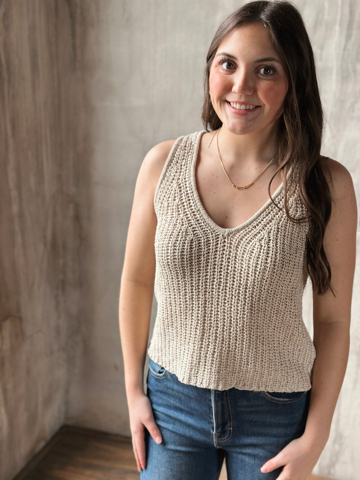 Woman wearing a beige knitted tank top and blue jeans against a neutral background