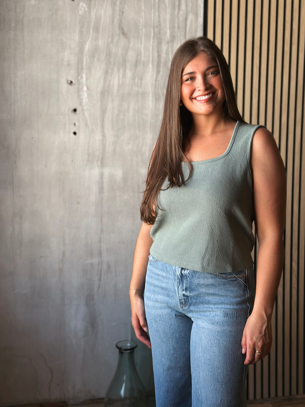 Woman wearing a light green sleeveless top and blue jeans standing against a textured wall.