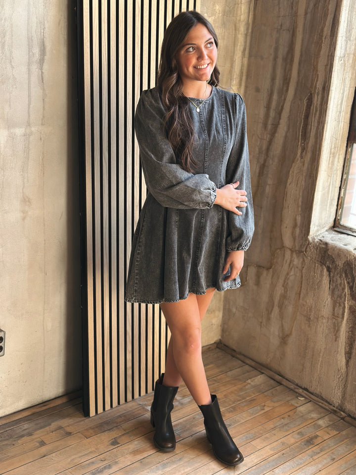 Woman in a gray dress standing against a wooden wall.