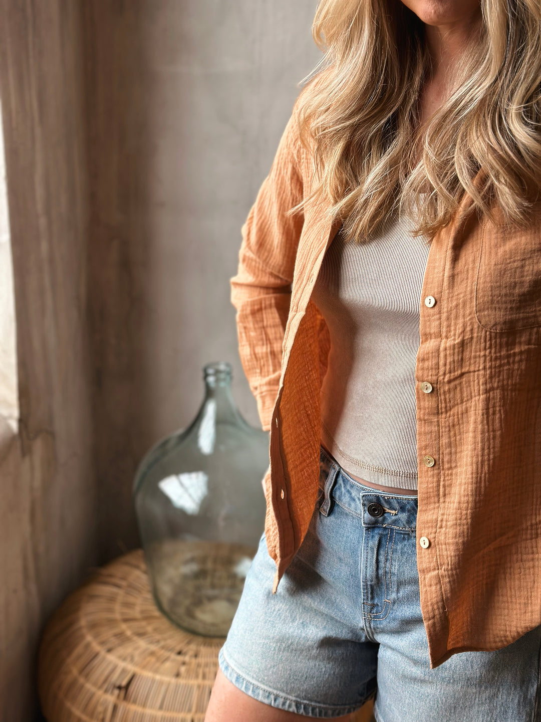 A person wearing a burnt orange blouse, white top, and denim shorts in a warm-toned room.