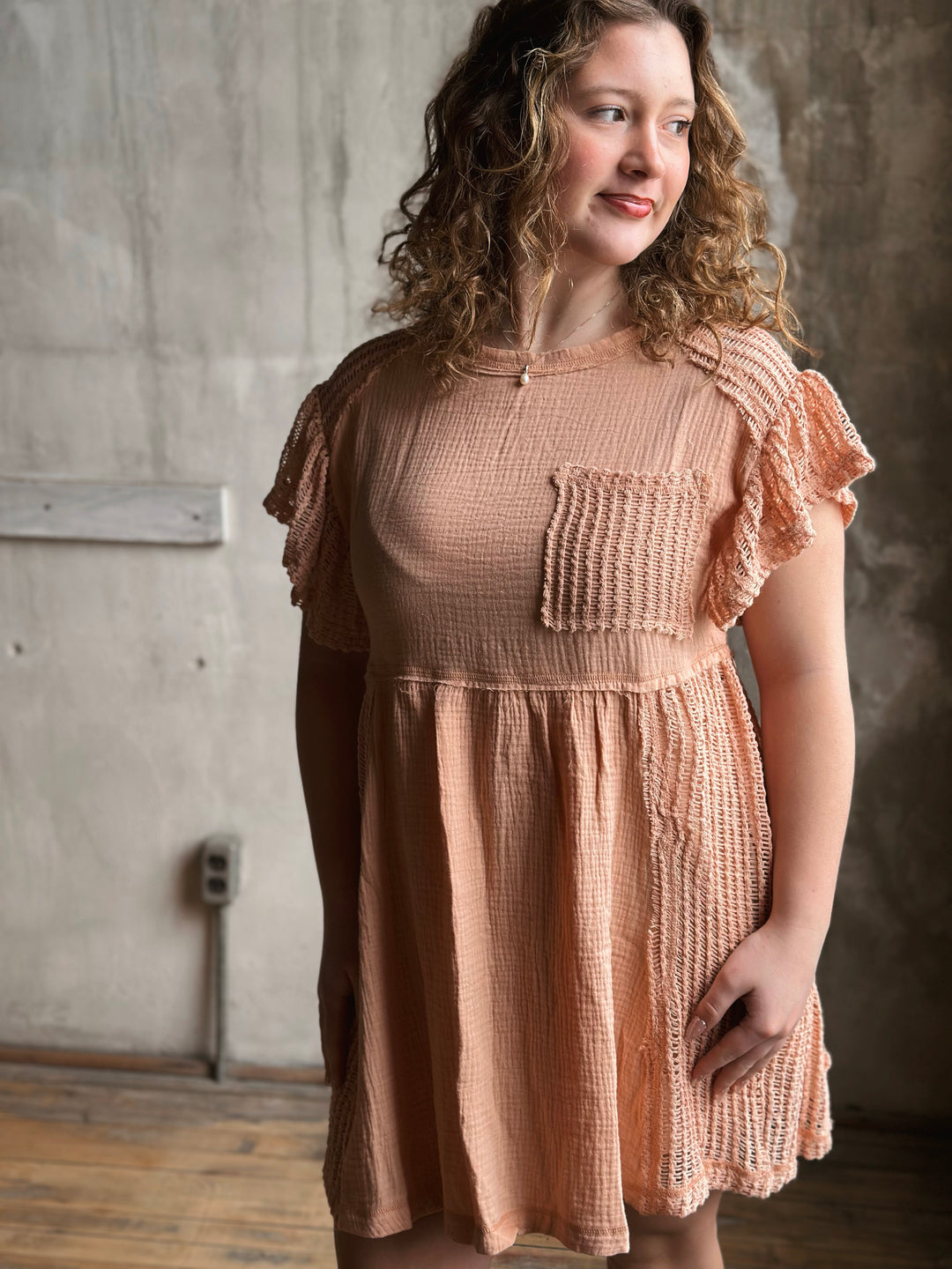 Woman wearing a peach-colored dress with ruffled sleeves in a room with concrete walls.