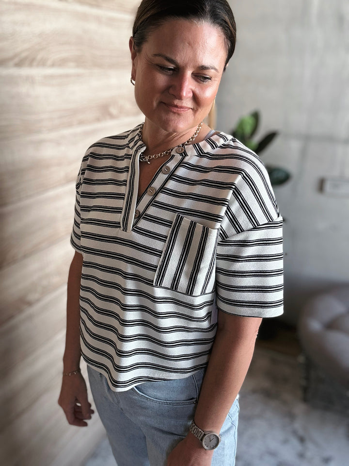 A woman wearing a short sleeved black and white striped v-neck shirt with buttons. With a pocket on the left side of the shirt. 