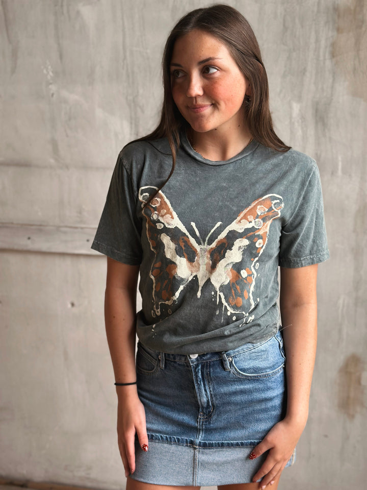 Woman wearing a gray t-shirt with a butterfly design against a concrete wall.
