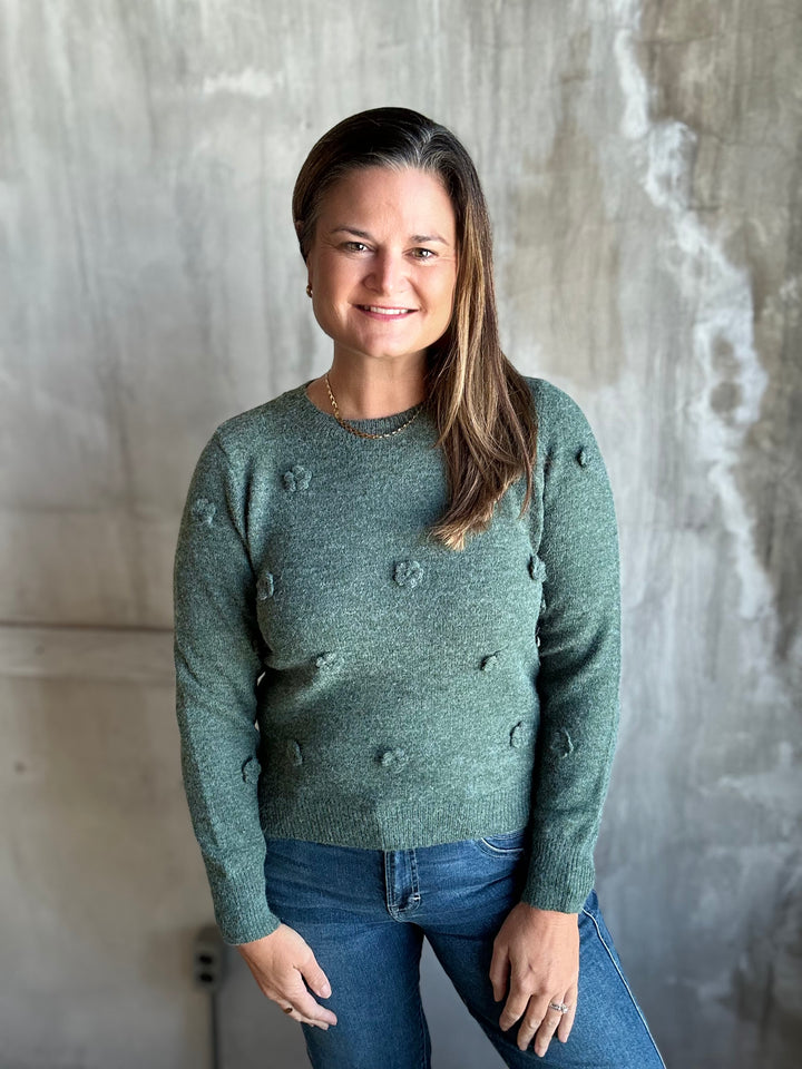 A woman with long brown hair wearing a textured sage green knit sweater and blue jeans, standing against a gray concrete wall.
