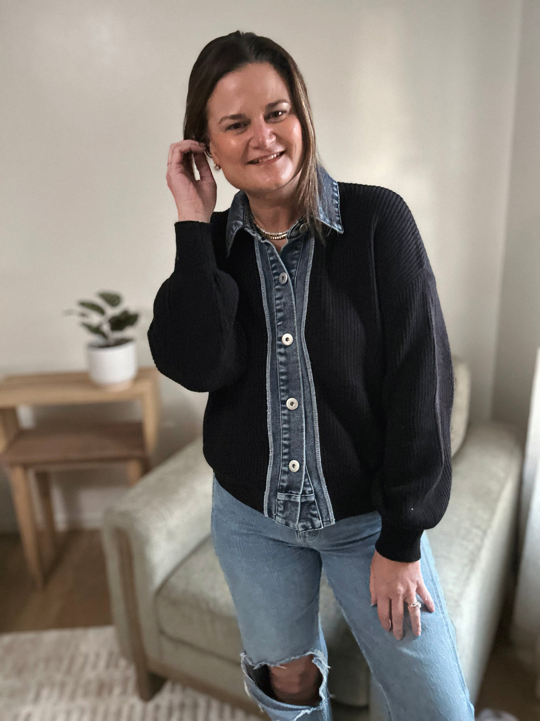 Woman wearing a black sweater over a denim shirt with ripped jeans indoors.