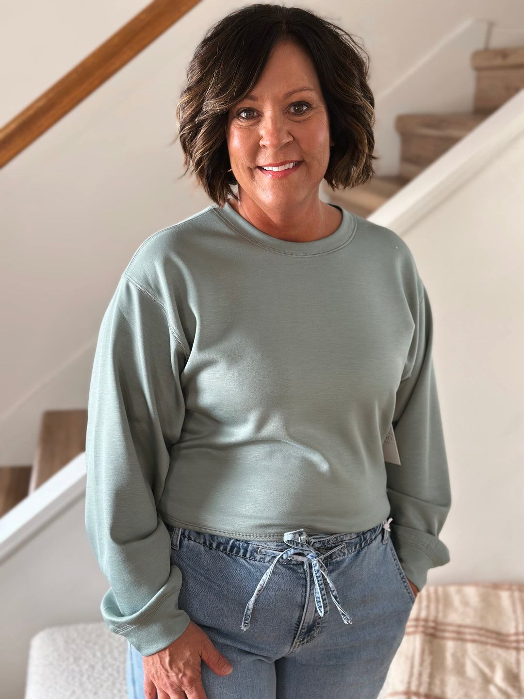 Woman wearing a light green sweatshirt and blue jeans standing indoors.