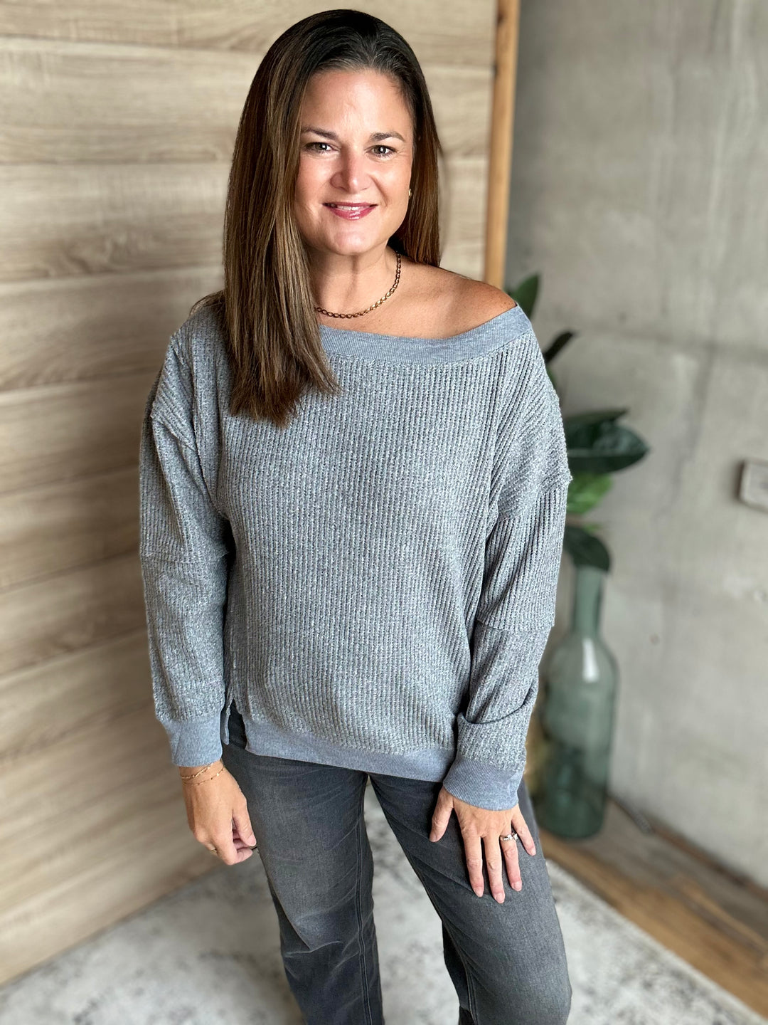 A woman wearing a heather gray off-the-shoulder ribbed knit sweater and dark gray jeans, standing in an indoor setting with a wood-paneled wall and a concrete wall in the background.
