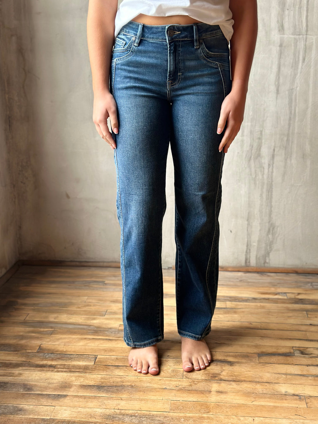 A person wearing medium-wash blue straight-leg jeans, standing barefoot on a wooden floor against a textured concrete wall.