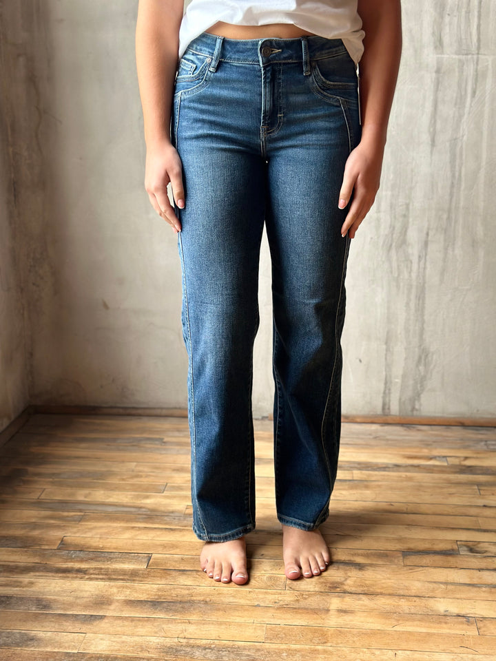 A person wearing medium-wash blue straight-leg jeans, standing barefoot on a wooden floor against a textured concrete wall.