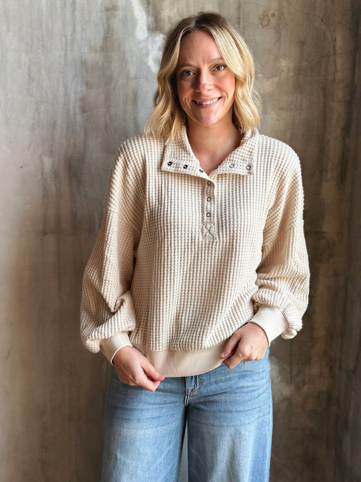 Woman wearing a beige textured sweater and blue jeans against a textured wall.