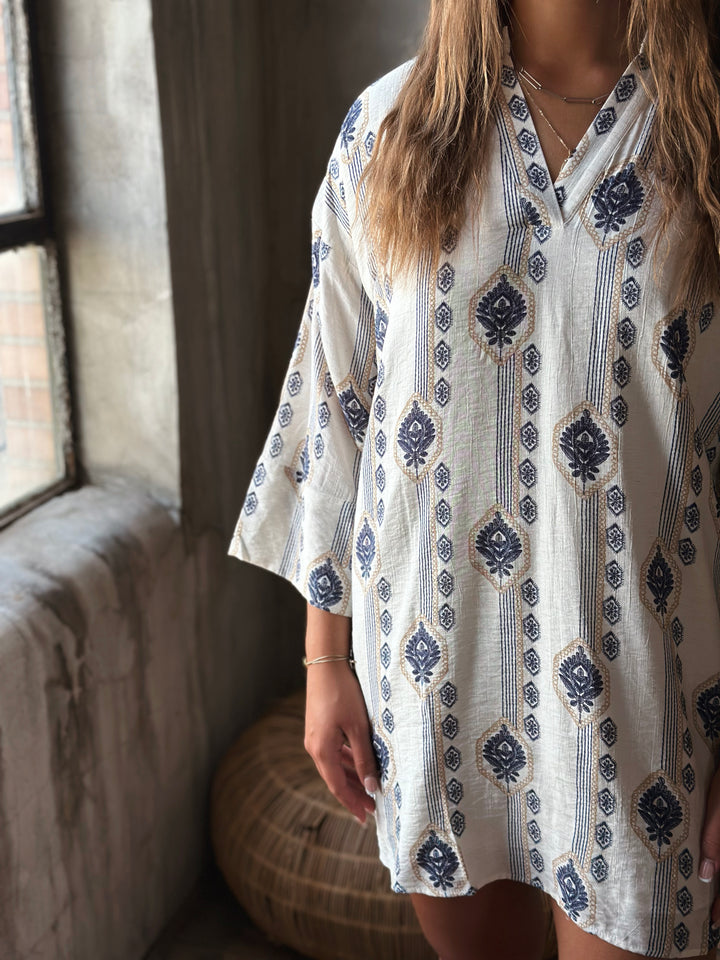 A person wearing a white v-neck dress with blue patterns in a room with a window and textured wall.