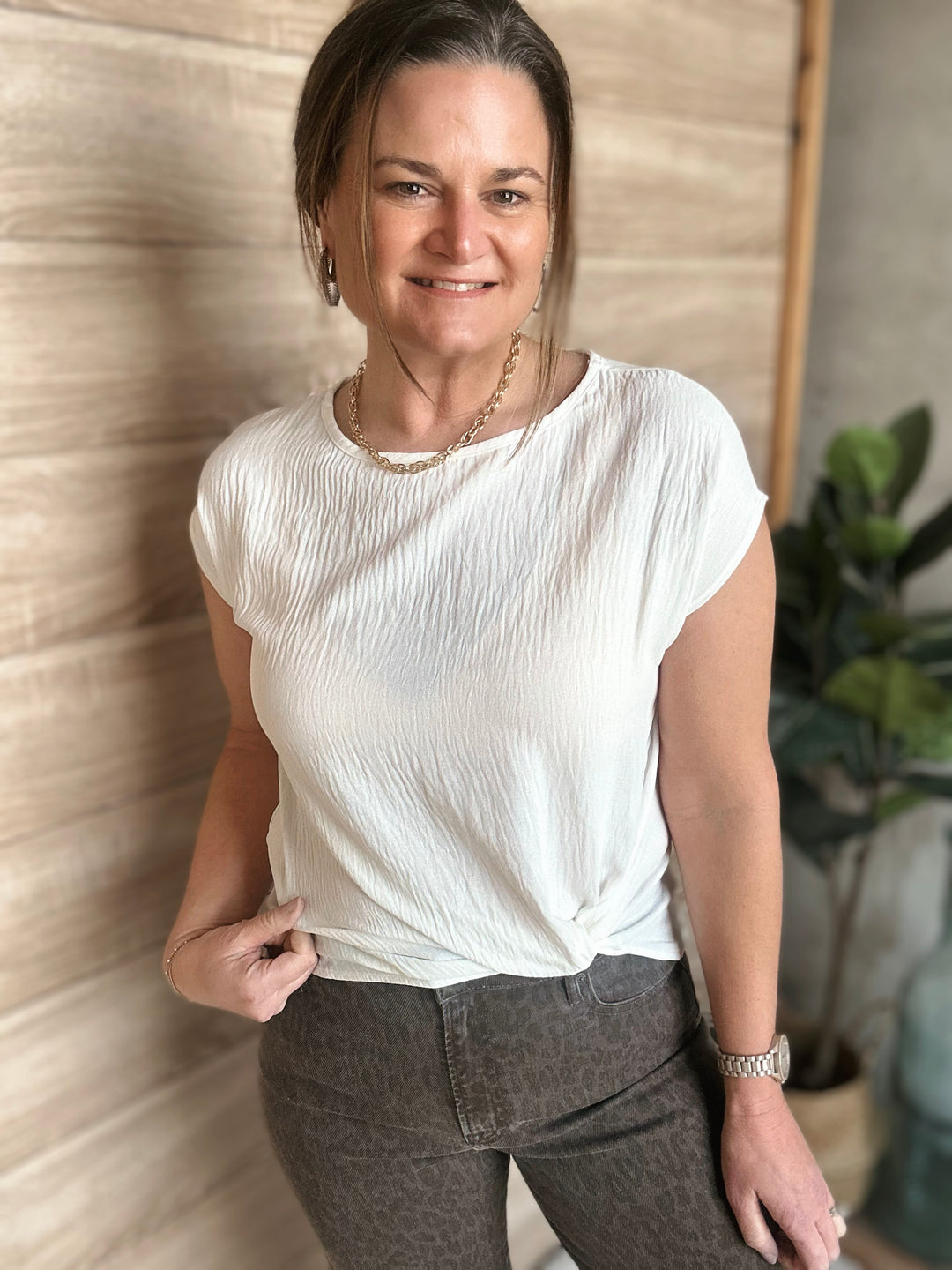 Woman wearing a white blouse and dark pants standing against a neutral wall with a plant in the background.