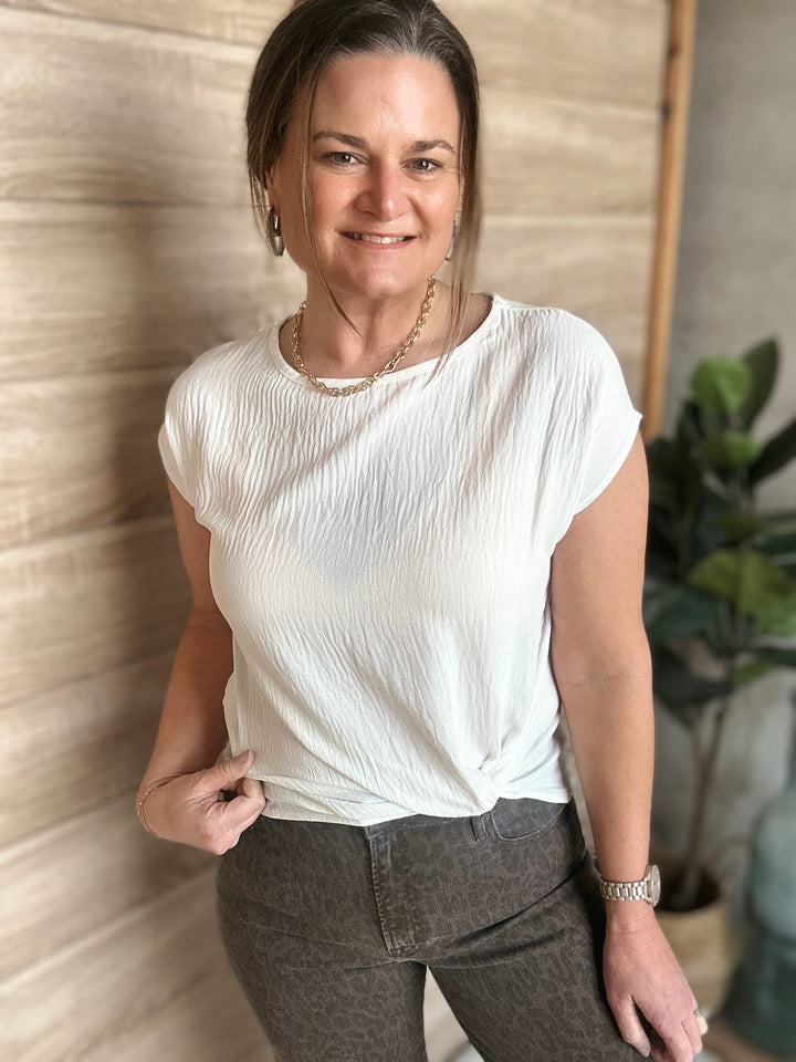 Woman wearing a white blouse and dark pants standing against a neutral wall with a plant in the background.
