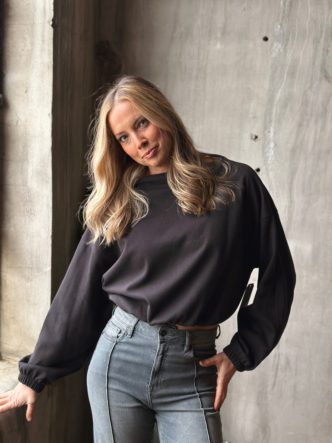 Woman wearing a black sweater and light blue jeans against a concrete wall.