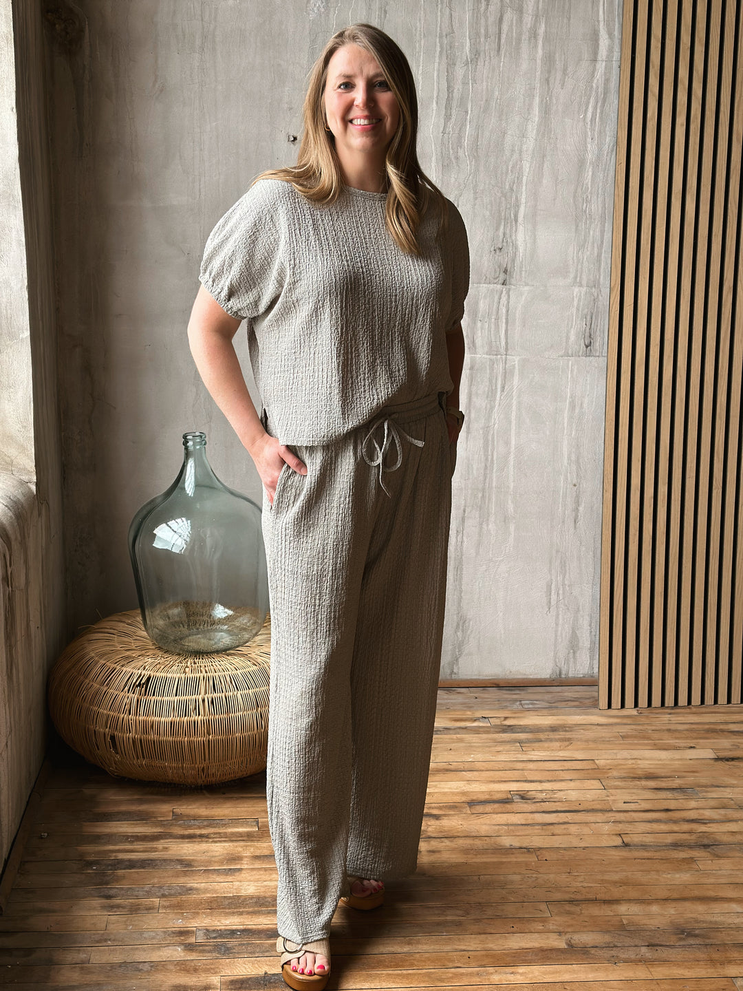 Woman wearing a matching sage outfit with a textured top and wide-leg pants in a room with wooden flooring and a large vase.