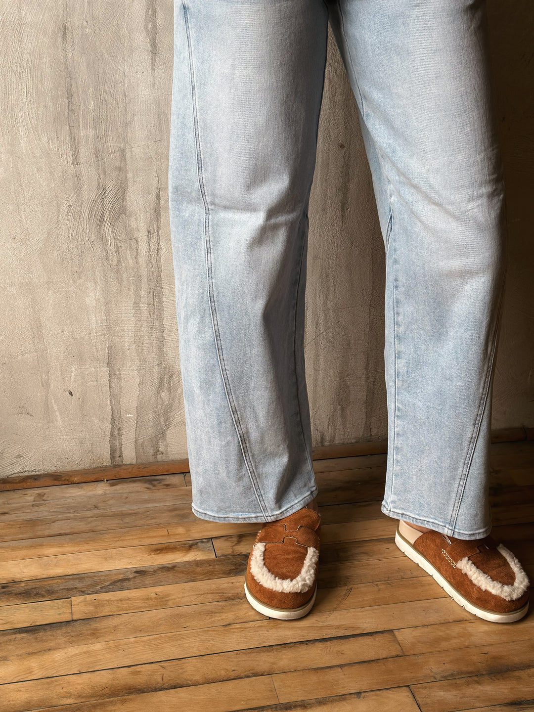 Person wearing light blue jeans and brown shoes on a wooden floor with a neutral wall background