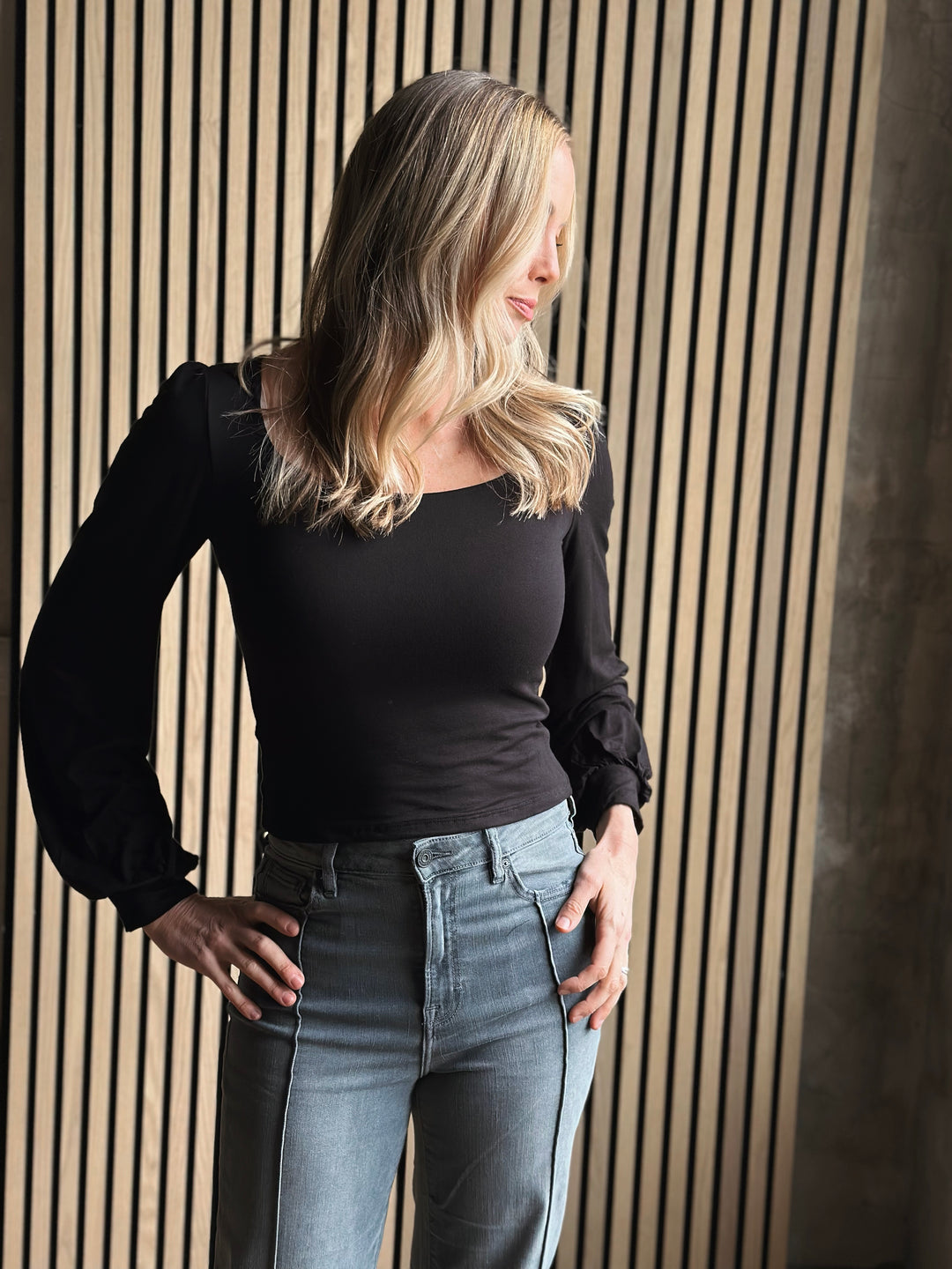 Woman wearing a black top and gray jeans against a wooden paneled wall.