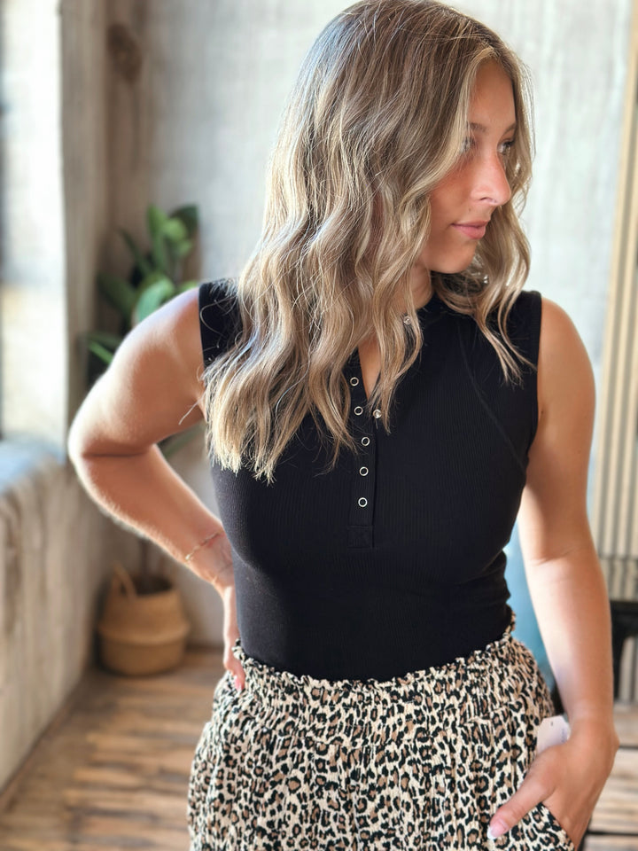 Woman wearing a black sleeveless top with button details and leopard print pants indoors.
