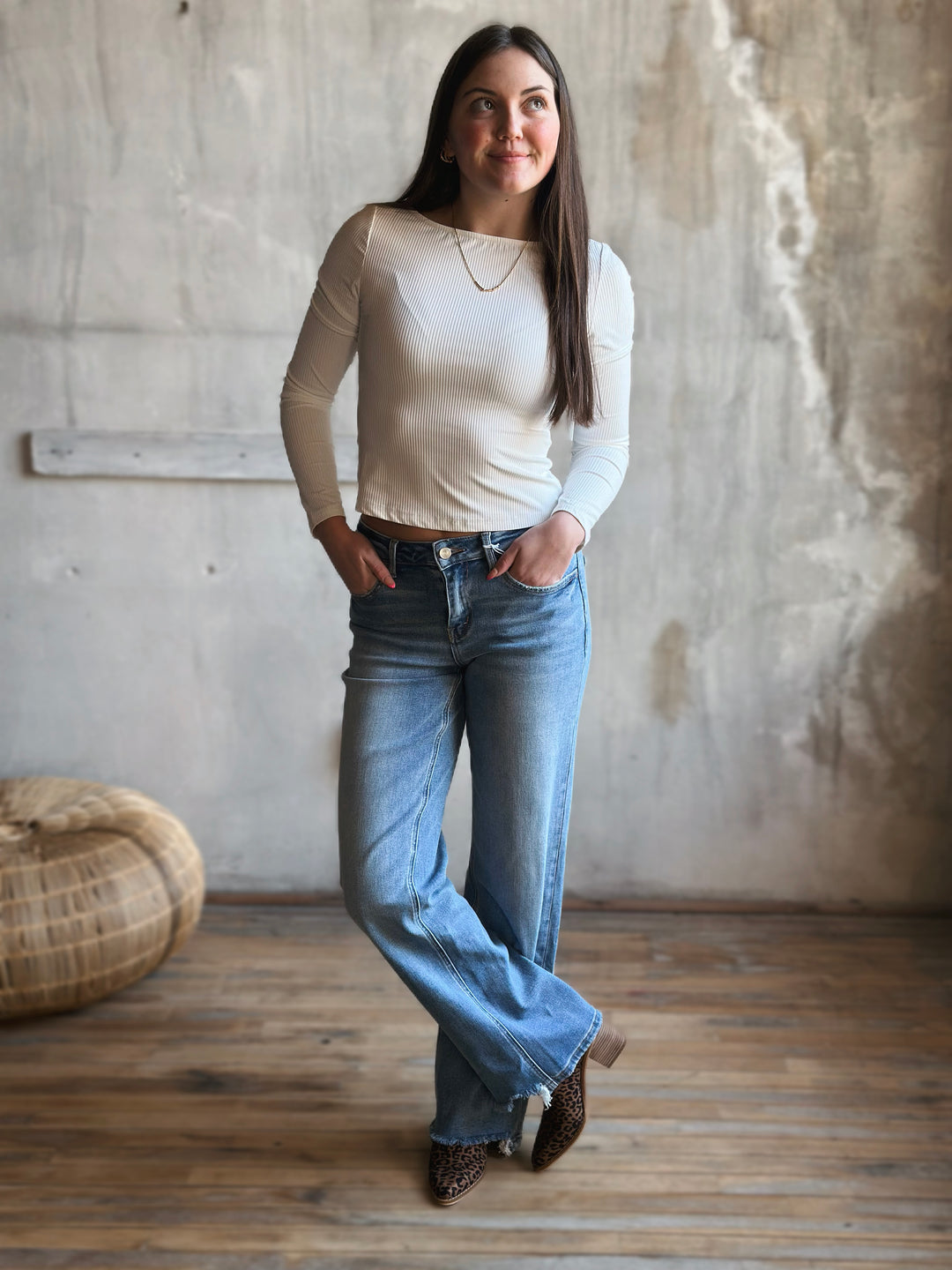 Woman wearing a white long-sleeve top and blue jeans standing in a room with wooden flooring and a textured wall.
