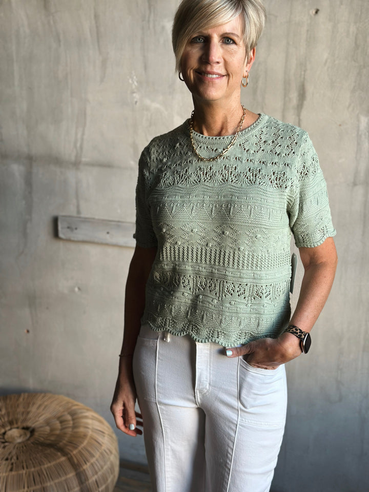 Woman wearing a green knitted top and white pants against a gray wall.