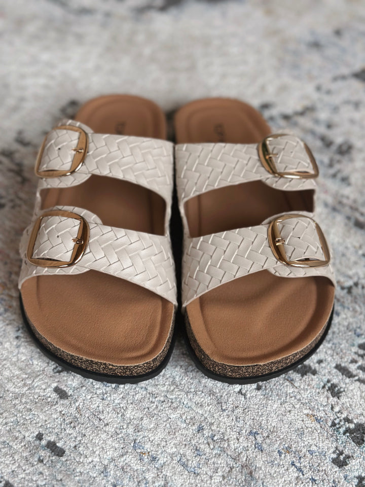 Pair of sandals with woven design and gold buckles on a textured surface.